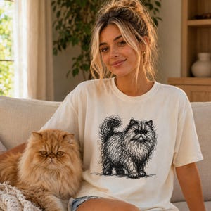 May include: A cream-colored short-sleeve t-shirt featuring a black ink-style sketch of a fluffy Persian cat. The graphic depicts the cat in a scribbled, artistic line-drawing technique, showcasing its long fur, rounded face, and bushy tail.