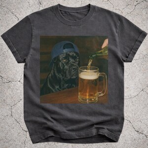 Könnte beinhalten: Dunkelgraues T-Shirt mit einem Aufdruck eines schwarzen Hundes mit einer blauen Baseballkappe, der einen Krug Bier betrachtet, der eingeschenkt wird. Der Hund befindet sich in einer Bar, mit einem Holztisch und dunklem Hintergrund.
