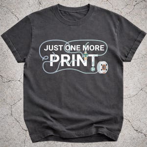Op de afbeelding: Donkergrijs t-shirt met de tekst "JUST ONE MORE PRINT" in het wit, omlijnd met lichtblauw. Het ontwerp bevat een 3D-printer en een spoel filament, wat wijst op een thema gerelateerd aan 3D-printen.