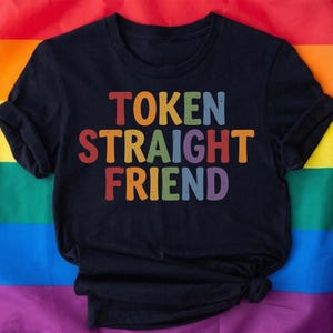 May include: Black t-shirt with the text "TOKEN STRAIGHT FRIEND" in colorful, block letters. The shirt is displayed against a rainbow flag background, representing the LGBTQ+ community.