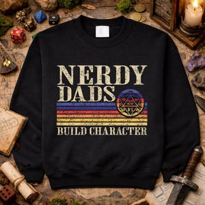 Könnte beinhalten: Schwarzes Sweatshirt mit dem Text "NERDY DADS BUILD CHARACTER". Das Design umfasst einen mehrfarbigen Streifen und eine Grafik eines 20-seitigen Würfels, ideal für Rollenspielfans.