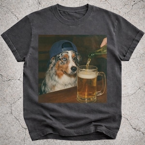 May include: Dark grey t-shirt with a graphic of a dog in a blue baseball cap, watching a beer mug being filled. The dog has blue eyes and a brown and white coat. The beer is a light amber colour.