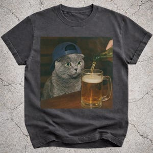 Op de afbeelding: Donkergrijs t-shirt met een afbeelding van een grijze kat met een blauwe baseballcap, die kijkt naar een bierpul die wordt gevuld met bier. De kat heeft groene ogen. De afbeelding staat tegen een bruine achtergrond.