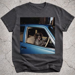 Può includere: Una t-shirt grigio antracite con una stampa grafica di un cane maculato marrone e bianco che indossa occhiali da sole neri, seduto al posto di guida di un'auto blu. Il cane ha le zampe sul volante e l'auto si trova in una città.