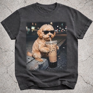 May include: Dark gray t-shirt featuring a cute dog wearing sunglasses and drinking iced coffee. The dog is sitting on a curb with a city background. The image is printed on a soft, comfortable t-shirt.