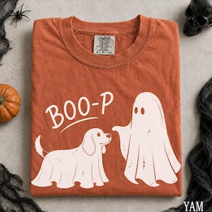 Peut inclure: Un t-shirt orange brûlé avec un motif blanc représentant un chien et un fantôme portant des draps, accompagné du texte BOO-P imprimé au-dessus du chien. Le t-shirt présente un col rond et une étiquette de marque Comfort Colors au col.