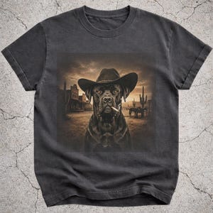 May include: A charcoal grey short-sleeve t-shirt featuring a graphic print of a black dog wearing a cowboy hat with a cigar in its mouth. The background of the print depicts a sepia-toned western desert landscape with buildings and cacti.