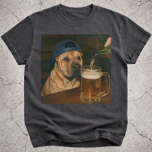 May include: Dark gray t-shirt featuring a Shar Pei dog wearing a blue baseball cap, looking at a beer mug being filled with beer. The image has a humorous theme, suggesting a relaxed, casual style.