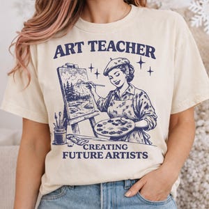 Puede incluir: Camiseta color crema con el texto "ART TEACHER" sobre una ilustración azul de una mujer pintando un paisaje. El texto "CREATING FUTURE ARTISTS" está debajo. La camiseta está hecha de un material suave.