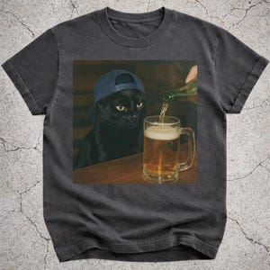 May include: Dark grey t-shirt with a humorous image of a black cat wearing a blue baseball cap, watching a beer mug being filled. The cat is central, with the beer mug and pouring hand in the background. The shirt has a vintage, worn appearance.