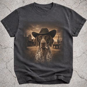Puede incluir: Una camiseta gris carbón de manga corta con un estampado gráfico central de un perro moteado que lleva un sombrero de vaquero marrón y un cigarrillo en la boca, sobre un paisaje desértico en tonos sepia con cactus y edificios antiguos.