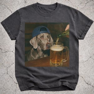 Peut inclure: T-shirt gris foncé avec une image imprimée d'un chien Weimaraner portant une casquette bleue, regardant de la bière être versée dans un verre. Le chien est à une table en bois. L'image est imprimée sur le devant du t-shirt.