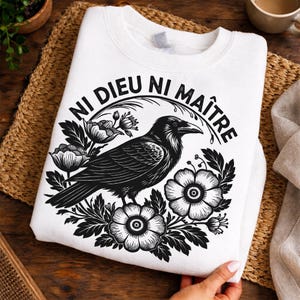 Puede incluir: Sudadera blanca con un diseño de cuervo negro, detalles florales y el texto "NI DIEU NI MAÎTRE". El diseño está impreso en una sudadera blanca de cuello redondo. El diseño presenta un cuervo y elementos florales.