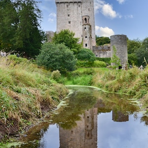 May include: A scenic view of a stone castle with a tall tower and a smaller round tower. The castle is reflected in the calm water of a stream in the foreground. Lush green vegetation surrounds the castle.