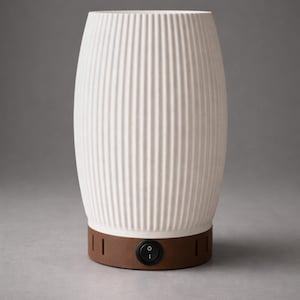 Puede incluir: Lámpara de cerámica blanca acanalada con base marrón. La lámpara tiene forma cilíndrica con ranuras verticales. Un interruptor de encendido/apagado negro está ubicado en la base marrón. La lámpara está diseñada para uso en interiores.