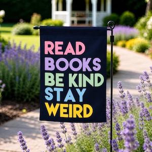 May include: A black garden flag with the words "READ BOOKS BE KIND STAY WEIRD" in colorful letters. The flag is displayed outdoors, surrounded by lavender plants and a pathway.
