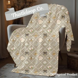 May include: A beige throw blanket featuring a repeating pattern of bees, envelopes, bows, candles, and flowers. The blanket is draped over a chair. The text "Thistleloop Co." is on a banner in the upper left corner.