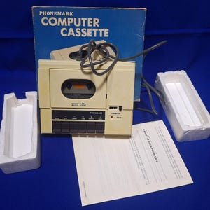 Könnte beinhalten: Ein beigefarbenes Phonemark Computer-Kassettendeck mit grauem Stromkabel, ausgestattet mit Kassettenschacht, Auto-Stop-Funktion und Bedientasten. Enthält einen blauen Originalkarton, weiße Styropor-Verpackungseinsätze und ein Anleitungsblatt.