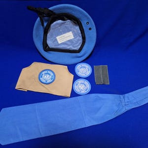 Pode incluir: Um boina azul claro, um patch de ombro bege, dois patches circulares e uma gravata azul são exibidos em uma superfície azul. Os patches e o patch de ombro apresentam um emblema azul e branco. A boina tem uma guarnição preta e um cordão preto.