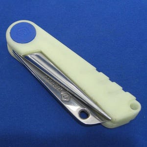 Peut inclure: Un outil pliable de couleur crème avec un bouton circulaire bleu et une lame argentée. La lame est partiellement déployée et porte l'inscription "Wichard". L'outil est sur fond bleu.