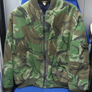 Op de afbeelding: Een camouflage bomberjack met een ritssluiting over de hele lengte, twee voorzakken en geribde manchetten en zoom. De jas heeft een groen, bruin en zwart patroon. De jas hangt aan een houten hanger.