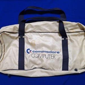 May include: A beige duffel bag with dark blue nylon straps and piping. The front features the blue Commodore logo and the word COMPUTER in black text. The bag has a zippered main compartment and metal rivets securing the handles to the body.