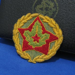 Könnte beinhalten: Gestickter Aufnäher mit einem roten Stern- und Ahornblatt-Design, umgeben von einem goldenen Lorbeerkranz. Der Aufnäher hat ein grünes und rotes Farbschema mit goldenen Akzenten. Wahrscheinlich ein militärischer oder Gedenkartikel.