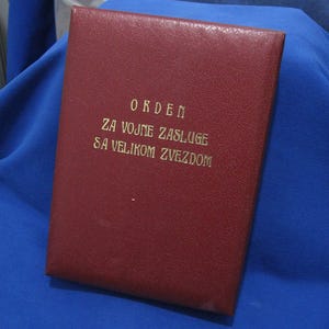 以下が含まれることがあります： 金色の文字が入った、バーガンディ色の長方形のケース。ケースのテキストは「ORDEN ZA VOJNE ZASLUGE SA VELIKOM ZVEZDOM」と書かれています。ケースは青い表面に置かれています。