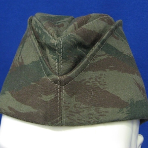 Pode incluir: Um chapéu com padrão de camuflagem em tons de verde e castanho. O chapéu tem formato triangular com aba dobrada e costura central. É feito de um tecido texturizado.