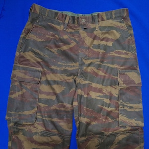 Puede incluir: Pantalones cargo de camuflaje con estampado de rayas de tigre. Los pantalones tienen cierre de botón, trabillas para el cinturón y dos bolsillos cargo en las piernas. La paleta de colores incluye tonos marrones, verdes y negros.