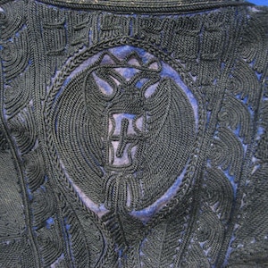 Puede incluir: Primer plano de un textil bordado en azul marino oscuro. El diseño presenta intrincadas puntadas en forma de cuerda que forman un emblema central con alas y una cruz, rodeado de patrones ornamentados. El fondo es azul claro.