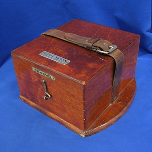 May include: A vintage wooden box with a rich, reddish-brown finish. It features a leather strap with a metal buckle and a small metal plaque. The box has a curved base and a metal latch. The text "ULYSSE NARDIN" is visible on the plaque.