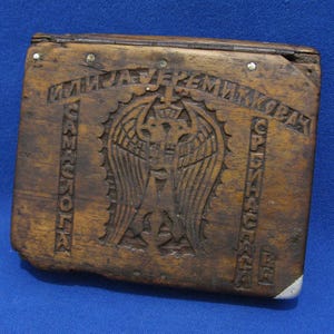 Op de afbeelding: Een rechthoekige houten doos met een scharnierend deksel met een uitgesneden embleem van een tweekoppige adelaar in het midden. Het hout heeft een donkerbruine afwerking met zichtbare nerven. Cyrillische tekst is in het oppervlak gegraveerd.
