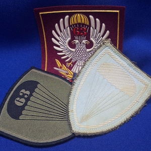 Op de afbeelding: Drie geborduurde patches met militaire ontwerpen. Eén is een rechthoekige bordeauxrode patch met een witte adelaar, een gele parachute en het nummer 63. Twee schildvormige patches zijn inbegrepen: één olijfgroen met een zwarte parachute en 63, en één lichtblauw met een witte parachute.