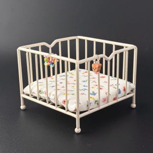 May include: A miniature cream-colored metal crib with a floral patterned pillow. The crib has vertical bars and rounded feet. Two small charms hang from the top of the crib, one with colorful beads and the other a small bear.