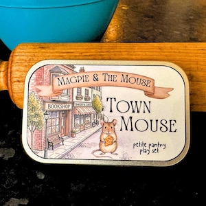 May include: A rectangular tin with the words "Magpie & The Mouse" and "Town Mouse" printed on it. The tin features an illustration of a town scene with a mouse holding cheese. A rolling pin and a blue bowl are in the background.
