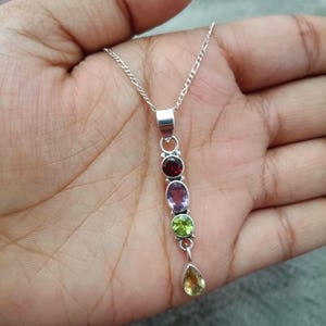 Può includere: Una collana d'argento con un pendente con una serie di pietre preziose colorate. Il pendente include una pietra rossa rotonda, una pietra viola ovale, una pietra verde rotonda e una pietra gialla a forma di goccia. La collana è esposta su uno sfondo neutro.