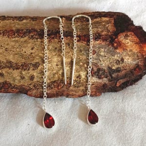 May include: A pair of silver chain earrings with a teardrop-shaped, deep red gemstone at the end of each chain. The earrings are displayed on a piece of textured wood.