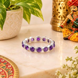 Pode incluir: Uma pulseira circular de prata com uma fileira contínua de pedras preciosas ametistas roxas, redondas e polidas, engastadas em engastes de metal individuais. A pulseira tem um design uniforme com cada pedra fixada numa moldura prateada.