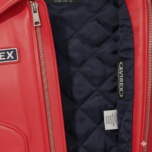 May include: Close-up of a red leather jacket with a silver zipper and a navy blue quilted lining. The jacket features a small patch with the word "EX" and a tag with the brand name "AVIREX".