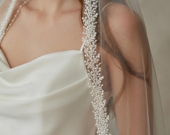 May include: A sheer white bridal veil featuring an intricate border of small white pearls arranged in a dense, branch-like pattern. The veil is draped over a smooth, sleeveless white dress with a draped cowl neckline and thin spaghetti straps.