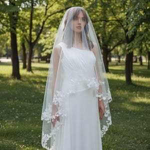 Op de afbeelding: Een bruid draagt een witte, off-shoulder trouwjurk en een doorschijnende sluier met bloemenkanten rand. De sluier valt over de voor- en achterkant en creëert een zachte, romantische uitstraling. De achtergrond is een vage groene park.