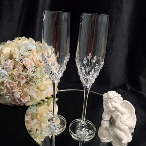 May include: Two clear champagne flutes with leaf and crystal accents. A bouquet of flowers and a small cherub statue are in the background. The flutes are on a mirrored surface.
