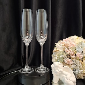 Op de afbeelding: Twee heldere champagneglazen met kristallen versieringen. De voet en het onderste deel van de glazen zijn versierd met glinsterende kristallen. Een boeket bloemen en een klein wit cherubijnbeeldje staan op de achtergrond. De glazen staan op een zwart oppervlak.