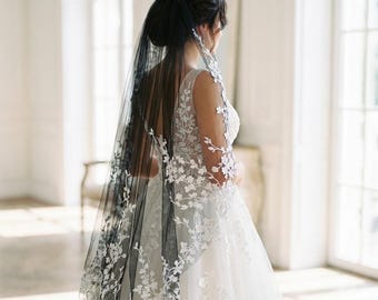 May include: A black tulle wedding veil featuring intricate white floral embroidery along the edges. The veil is attached to a hair comb and is worn over a white sleeveless gown with matching white floral lace detailing on the bodice and shoulders.