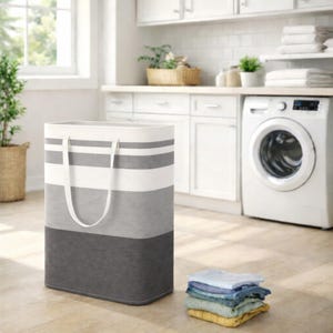 Puede incluir: Una cesta de lavandería rectangular con una parte superior blanca, rayas grises y blancas y una base gris oscuro. Tiene asas blancas y está junto a una pila de ropa doblada. La cesta está en un lavadero.