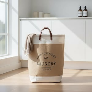 Puede incluir: Una cesta de lavandería beige y blanca con asas de cuero marrón. La cesta tiene las palabras "WASH. DRY. IRON. LAUNDRY DELUXE" impresas en la parte delantera. Una toalla beige cuelga de un lado. La cesta está en una habitación luminosa.