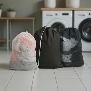 Puede incluir: Tres bolsas de lavandería en un lavadero. Una es una bolsa de malla blanca llena de ropa, otra es una bolsa negra sólida con un cordón y la tercera es una bolsa de malla negra. Las bolsas están sobre un suelo embaldosado.