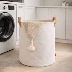 May include: A cylindrical, off-white laundry hamper with brown leather handles and a decorative tassel. The hamper has a neutral color palette and is made of a textured fabric. The hamper is approximately 24 inches tall and 18 inches in diameter.