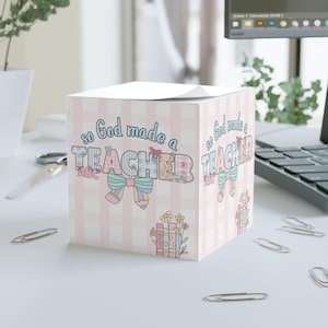 Peut inclure: Distributeur de blocs-notes carré avec un motif vichy rose et blanc. Le texte "so God made a TEACHER" est affiché en lettres colorées, avec un nœud et un crayon. Un motif floral et de livre est également présent.